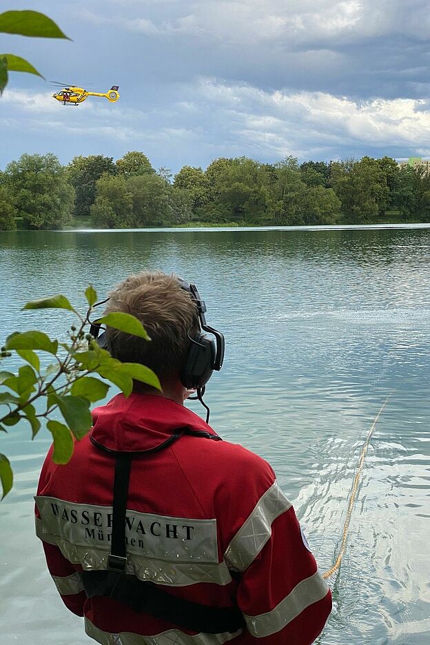 Ein Leinenführer der Wasserwacht mit einem Headset auf dem Kopf spricht mit einem Taucher, während ein gelber Hubschrauber des ADAC über dem See schwebt.